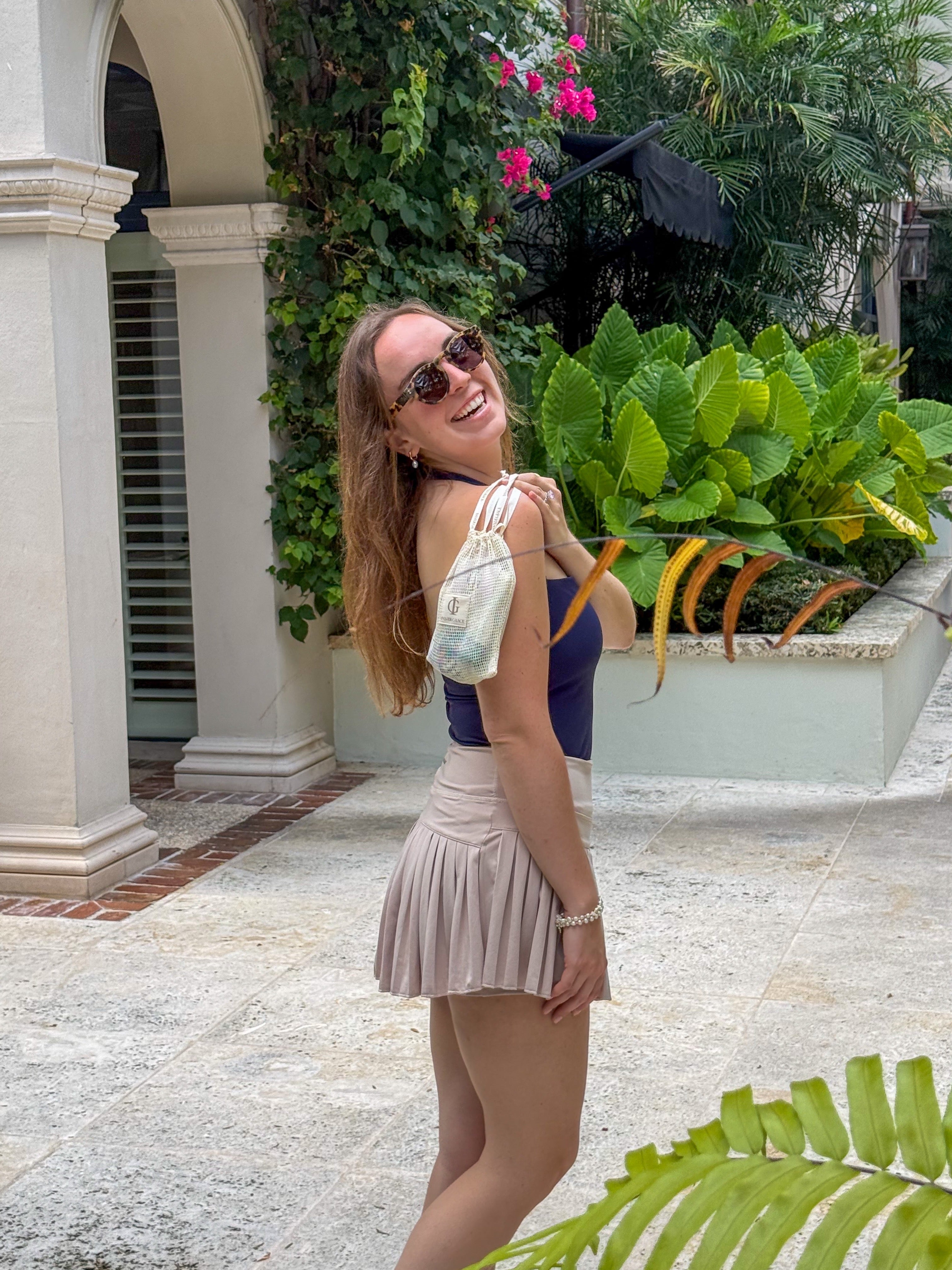 Woman in sunglasses and a purple top with a pleated skirt standing in a tropical setting.
