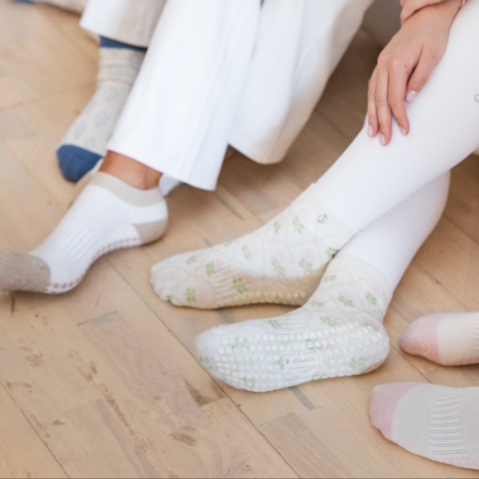 Three people sitting on a wooden floor wearing white and light pink tights wearing pilates socks